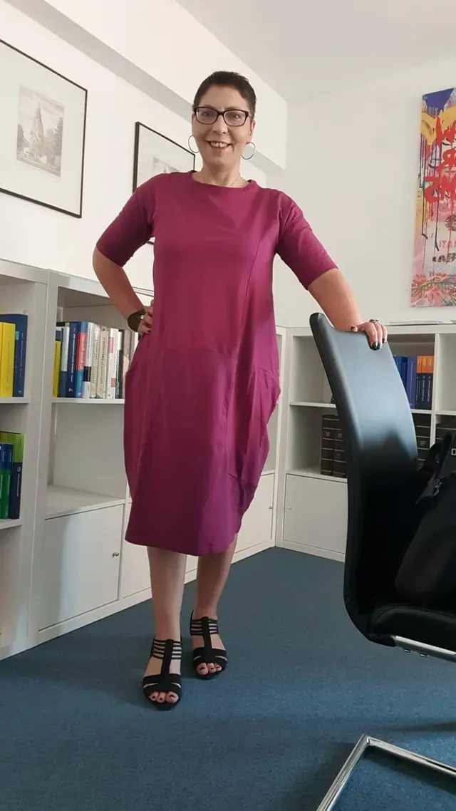 Sina Riegel steht lächelnd neben einem Bürostuhl vor Bücherregalen